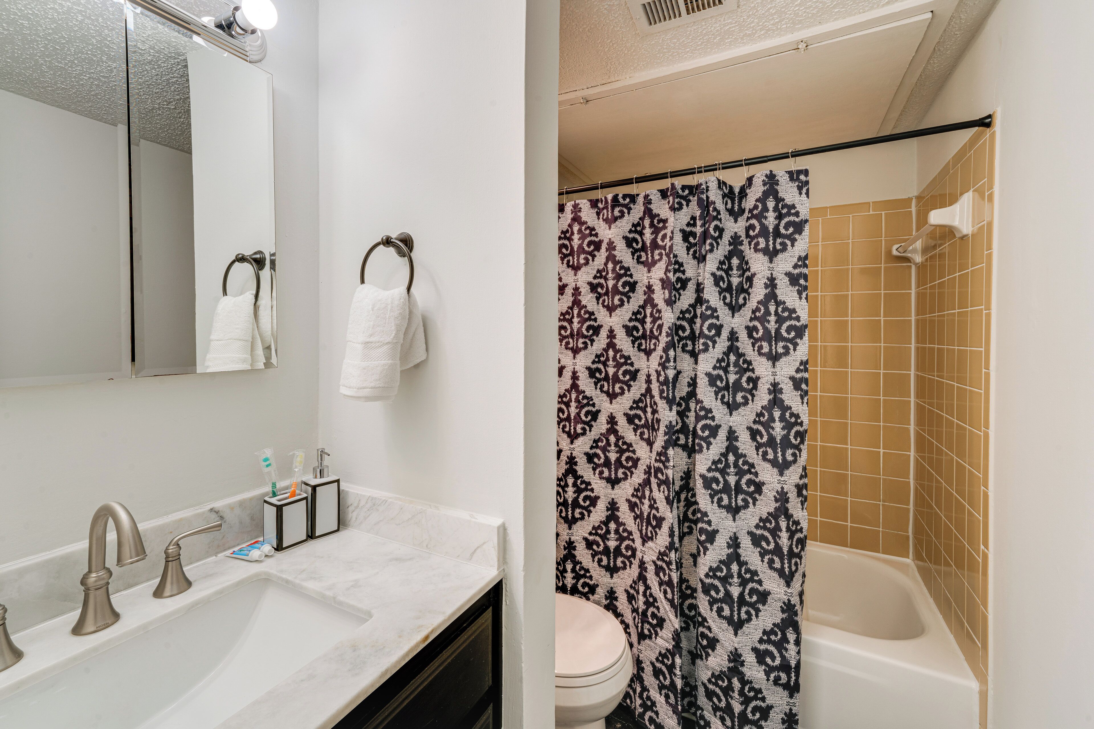A bathroom with a sink, three faucets, a mirror, a toilet, and a shower curtain with a patterned design.