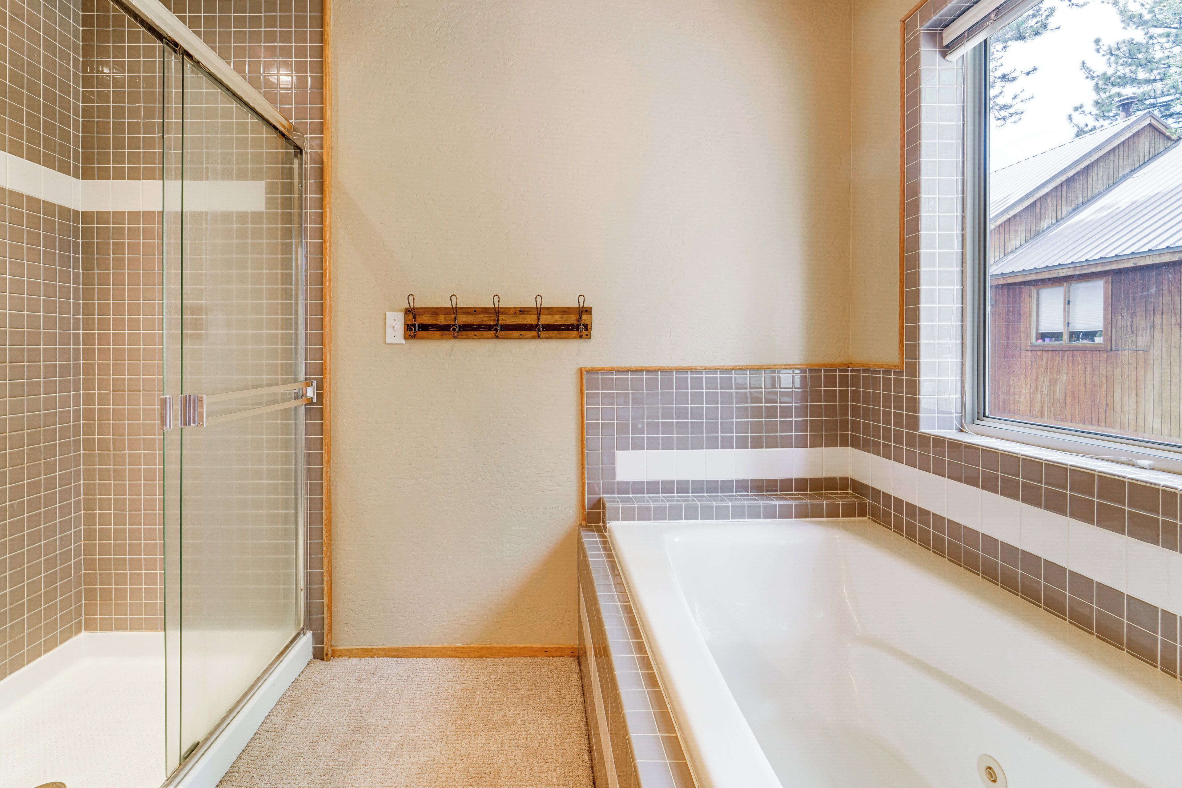 A bathroom with a glass shower enclosure, a bathtub, and a window providing a view of a wooden building outside.