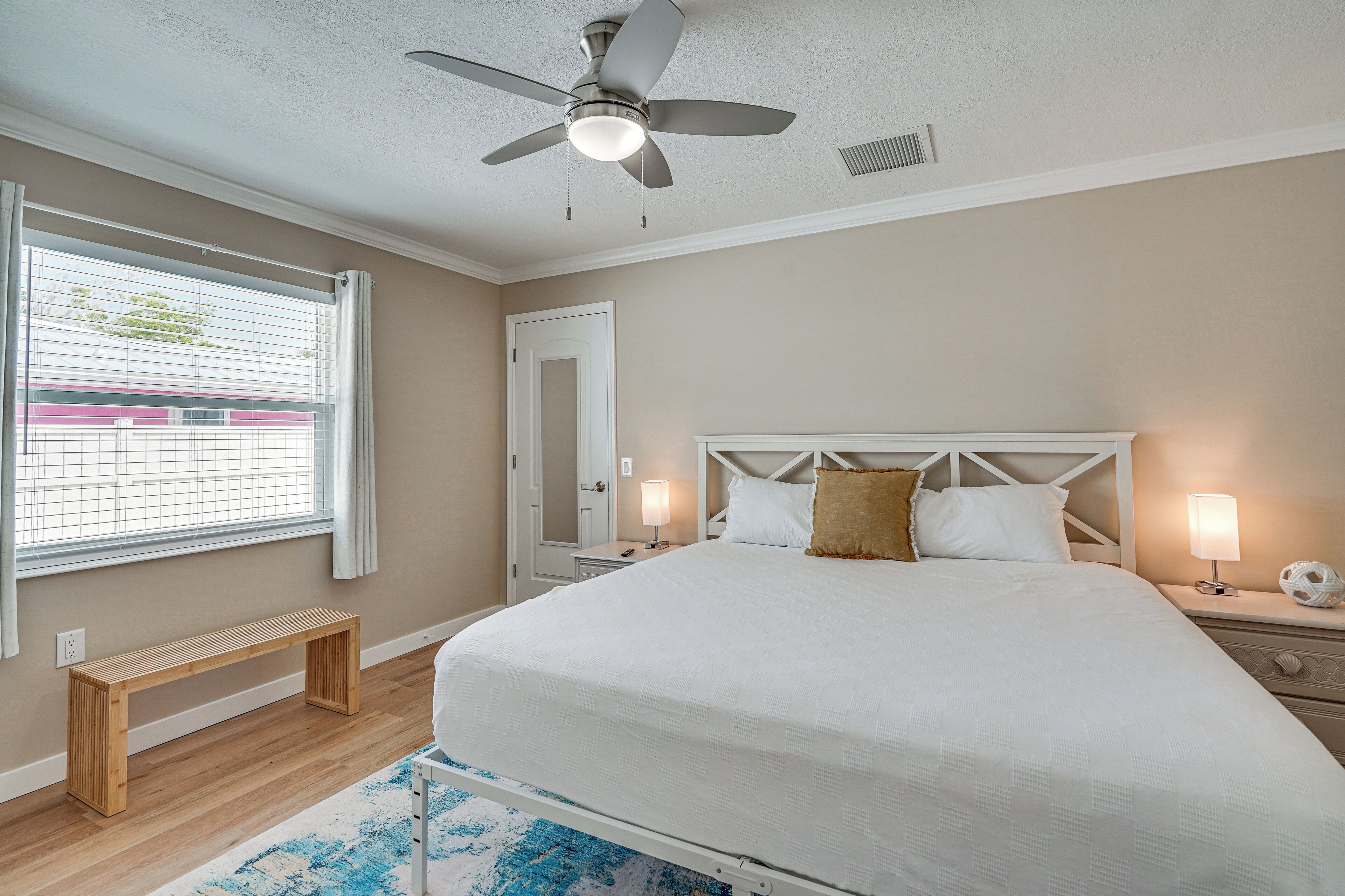 Une chambre à coucher avec un lit, des lampes de chevet, un ventilateur de plafond, un banc et une fenêtre avec des stores.