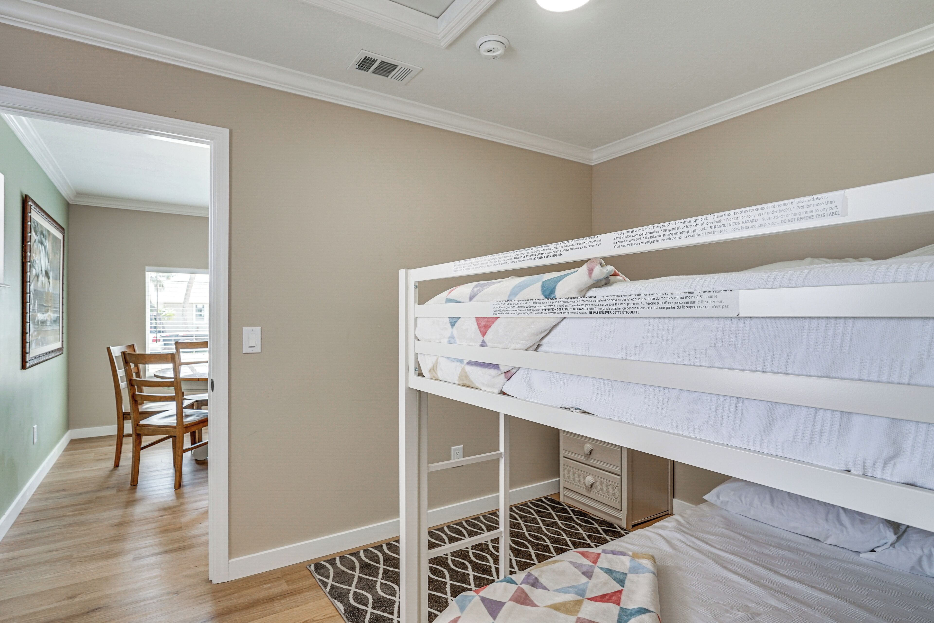 Una cama litera con un cajón para guardar cosas, una mesa de comedor de madera con sillas y una alfombra estampada.