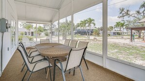 A covered patio with a wicker table and chairs, large windows, and a view of a residential area with palm trees.