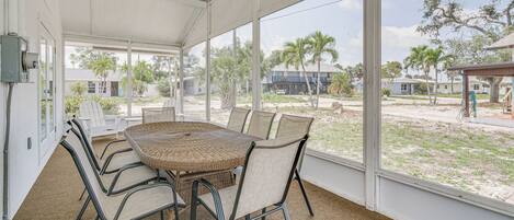 A covered patio with a wicker table and chairs, large windows, and a view of a residential area with palm trees.