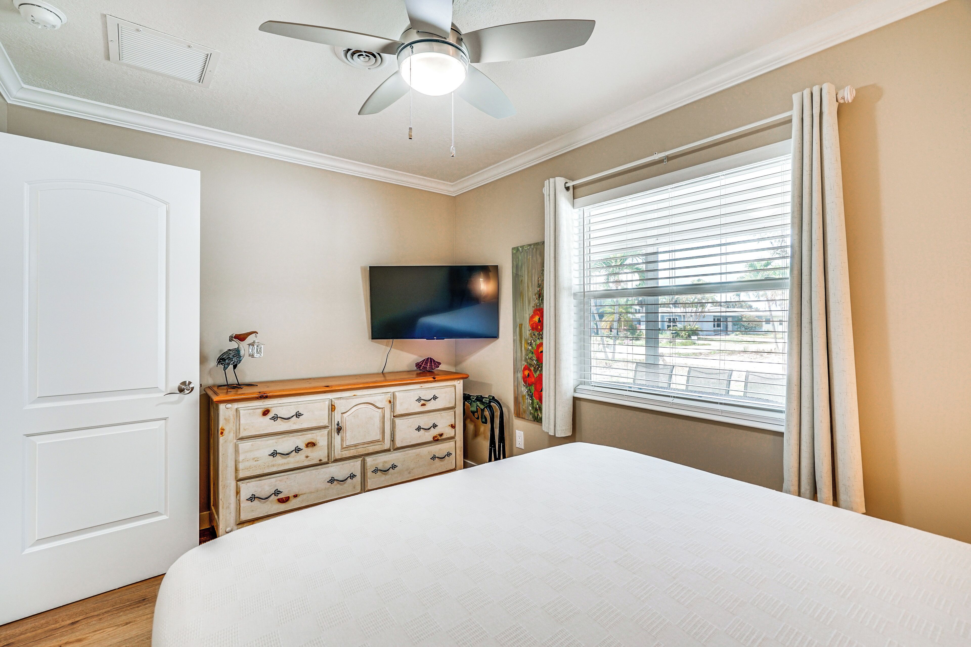 Un dormitorio con cama, cómoda, televisor, ventilador de techo, ventana con persianas y una puerta.