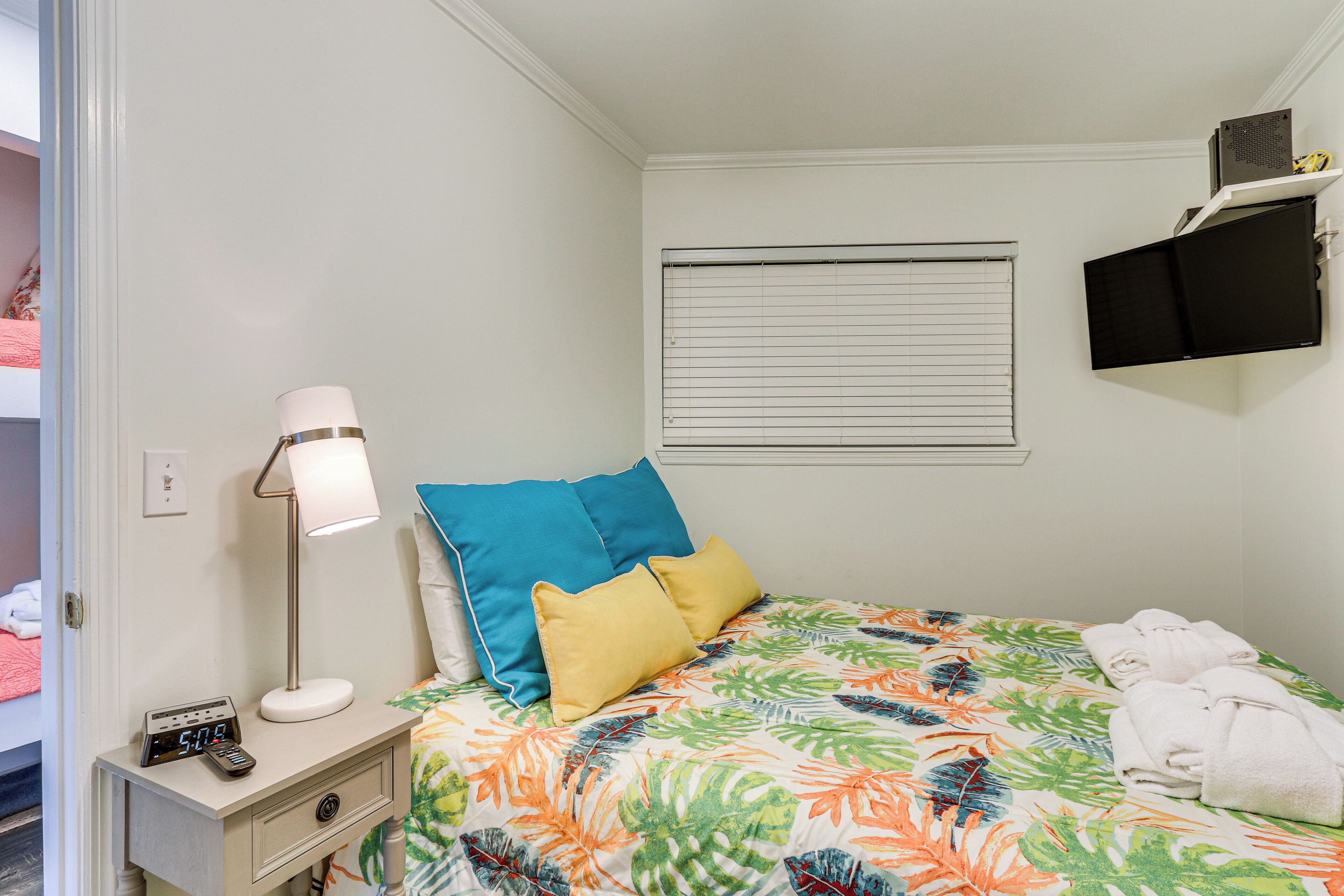 A neatly made bed with a tropical leaf pattern bedspread, a bedside lamp, and a digital clock. There is a window with blinds, a flat-screen TV mounted on the wall, and a small shelf with towels.