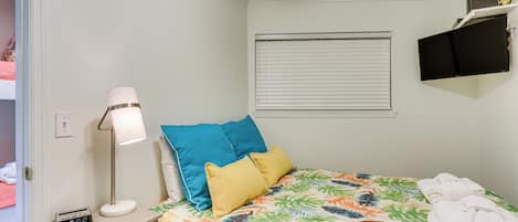 A neatly made bed with a tropical leaf pattern bedspread, a bedside lamp, and a digital clock. There is a window with blinds, a flat-screen TV mounted on the wall, and a small shelf with towels.