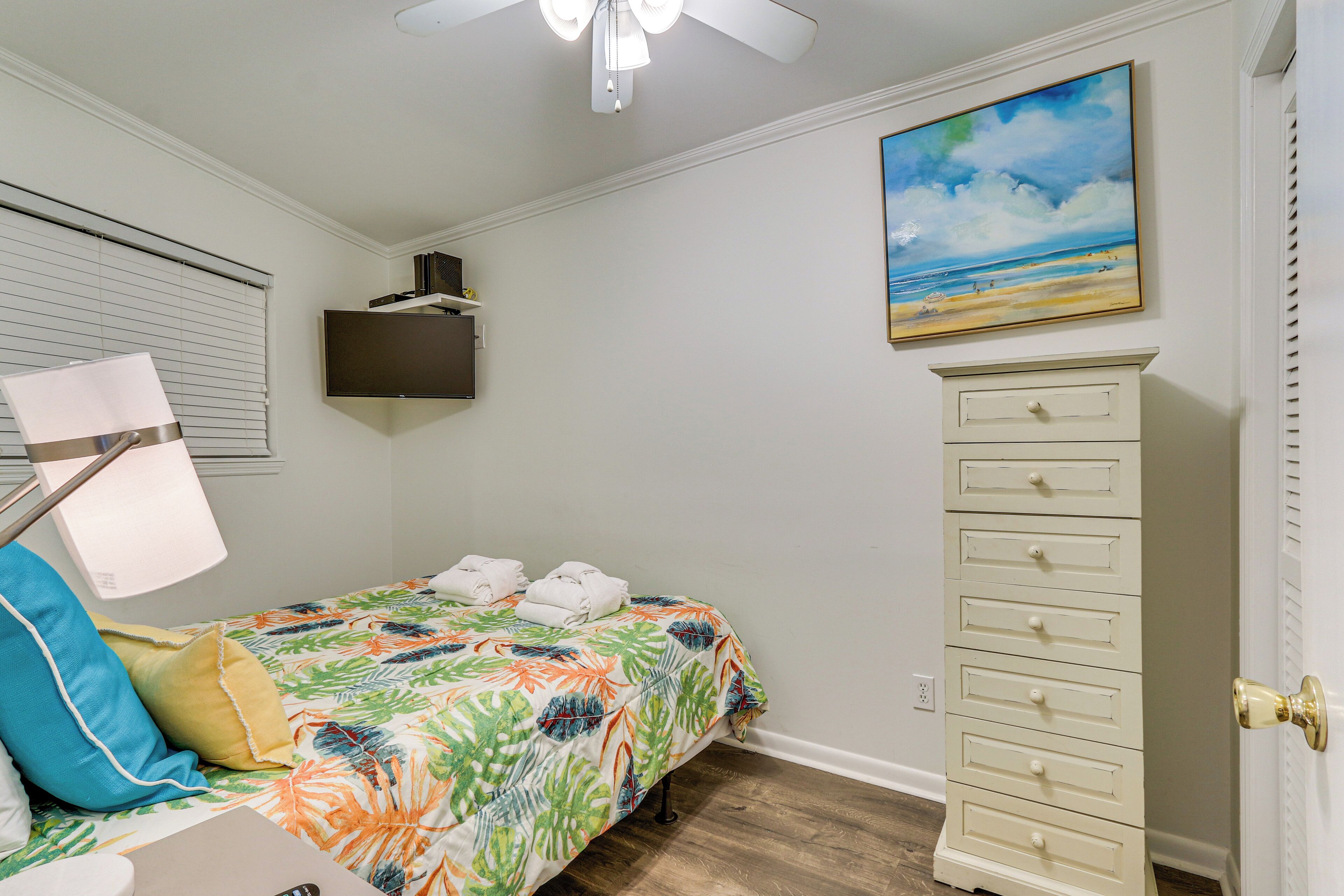 A bedroom with a bed, a dresser, a ceiling fan, a painting, and a TV on the wall.