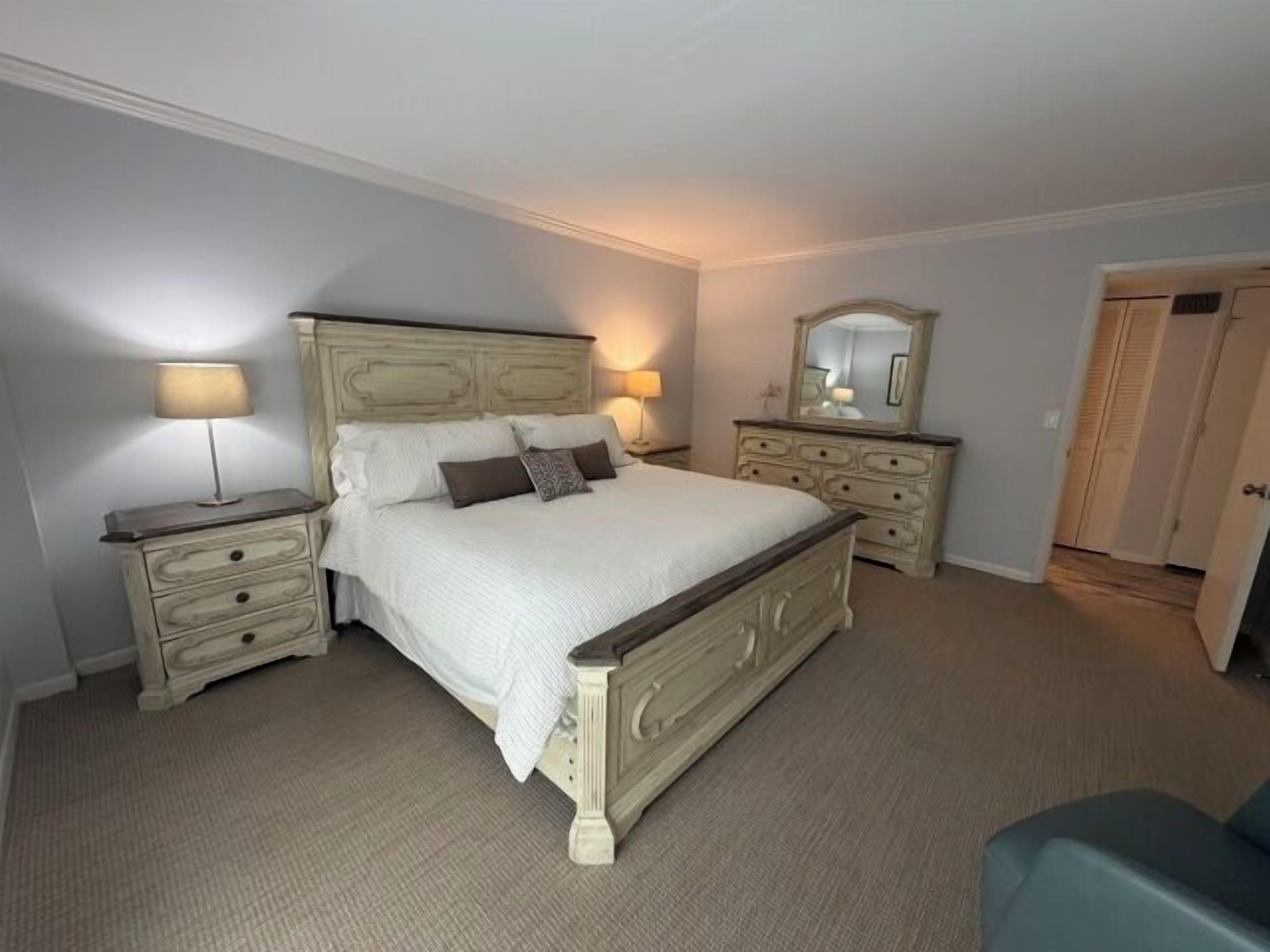 A bedroom with a large bed, two bedside tables, a dresser, a mirror, and a door.