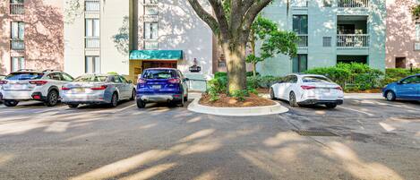 A parking lot with several cars parked in front of a building with balconies and a tree in the center.