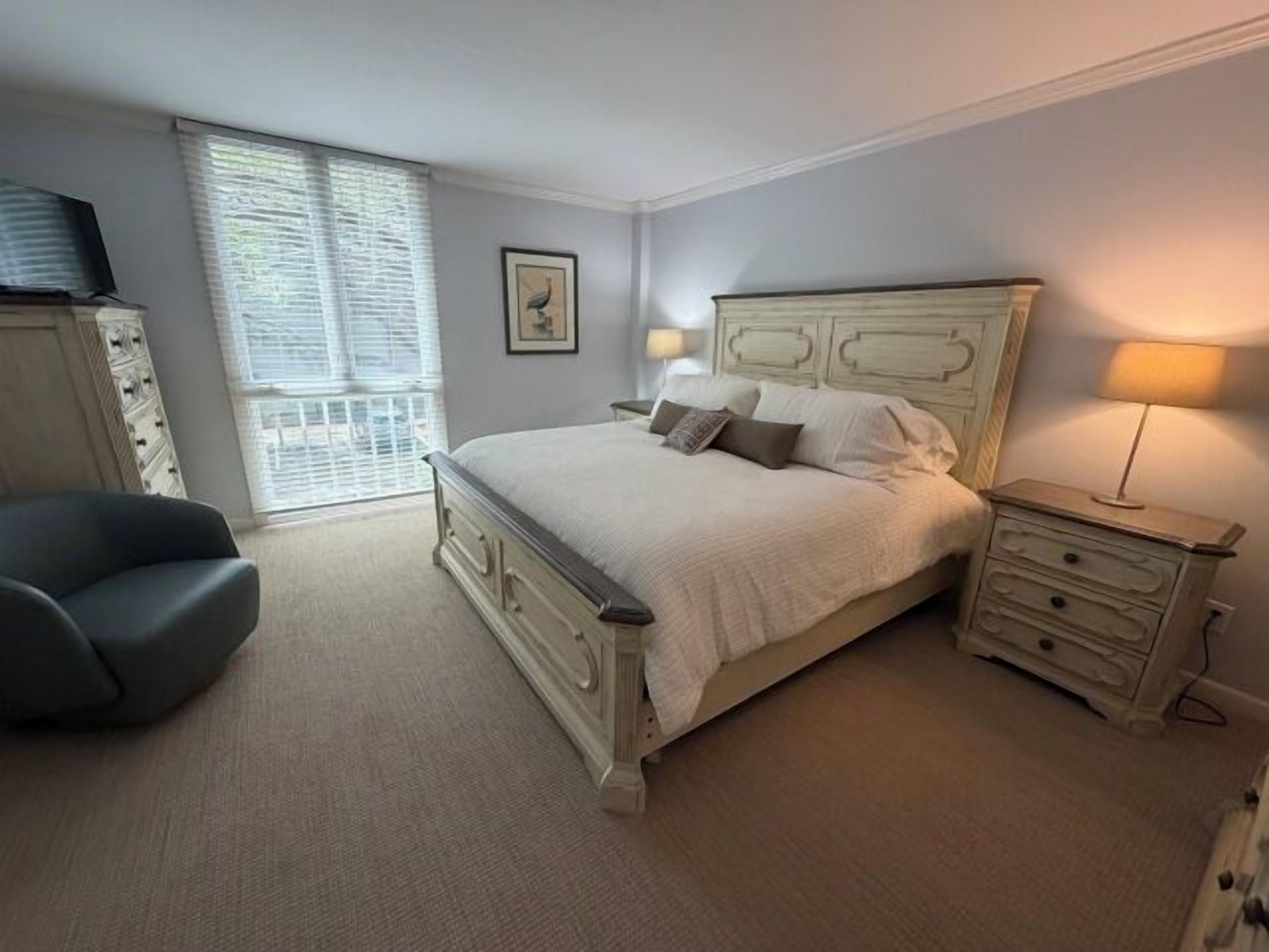 A bedroom with a bed, bedside tables, a chair, a window with blinds, and a picture on the wall.