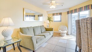 A living room with a sofa, a table with a lamp, a chair, a ceiling fan, and a large window with blinds. - Pool & Ocean View: Myrtle Beach Resort Condo! (Myrtle Beach)