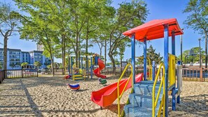 A playground with slides and climbing structures, surrounded by trees and a building in the background. - Pool & Ocean View: Myrtle Beach Resort Condo! (Myrtle Beach)