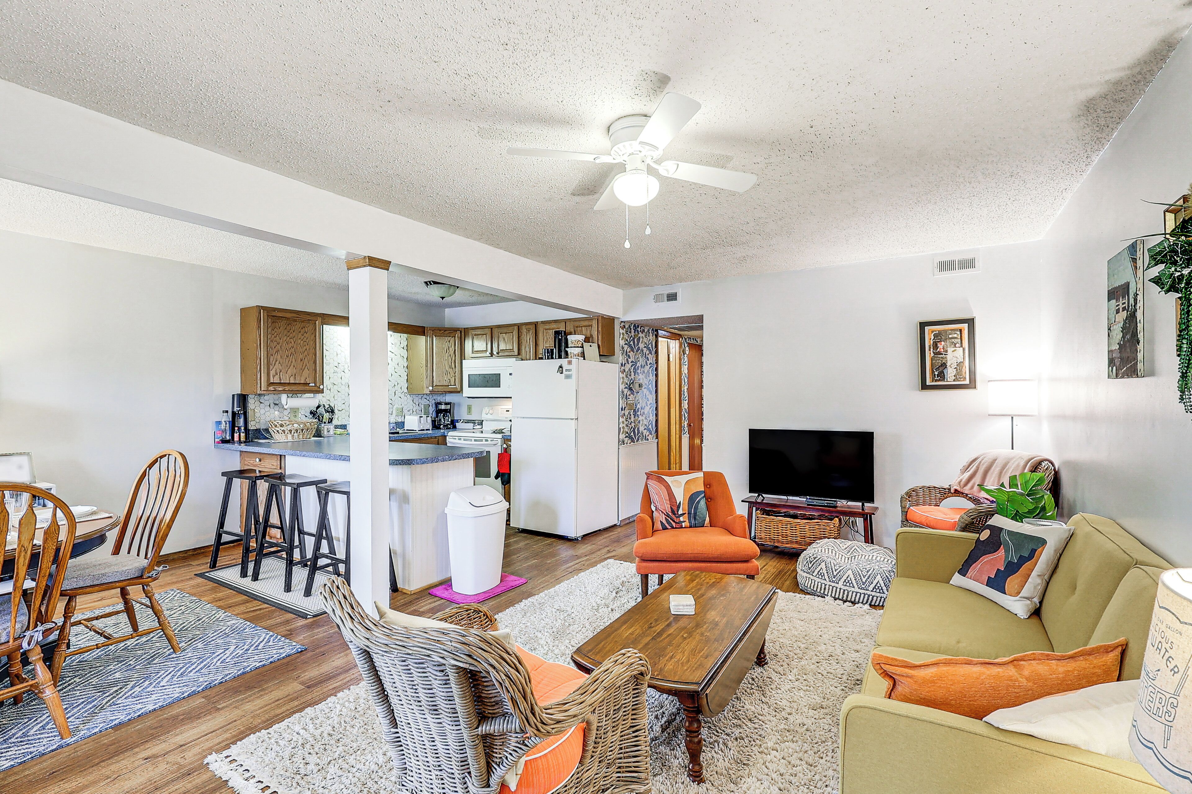 A living room with a sofa, chairs, a coffee table, and a TV. There is a kitchen in the background.