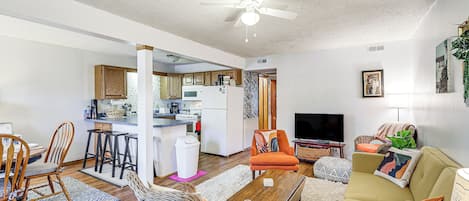 A living room with a sofa, chairs, a coffee table, and a TV. There is a kitchen in the background.