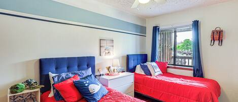 Une chambre avec deux lits, un ventilateur de plafond, une fenêtre avec des rideaux et une petite table avec une lampe.