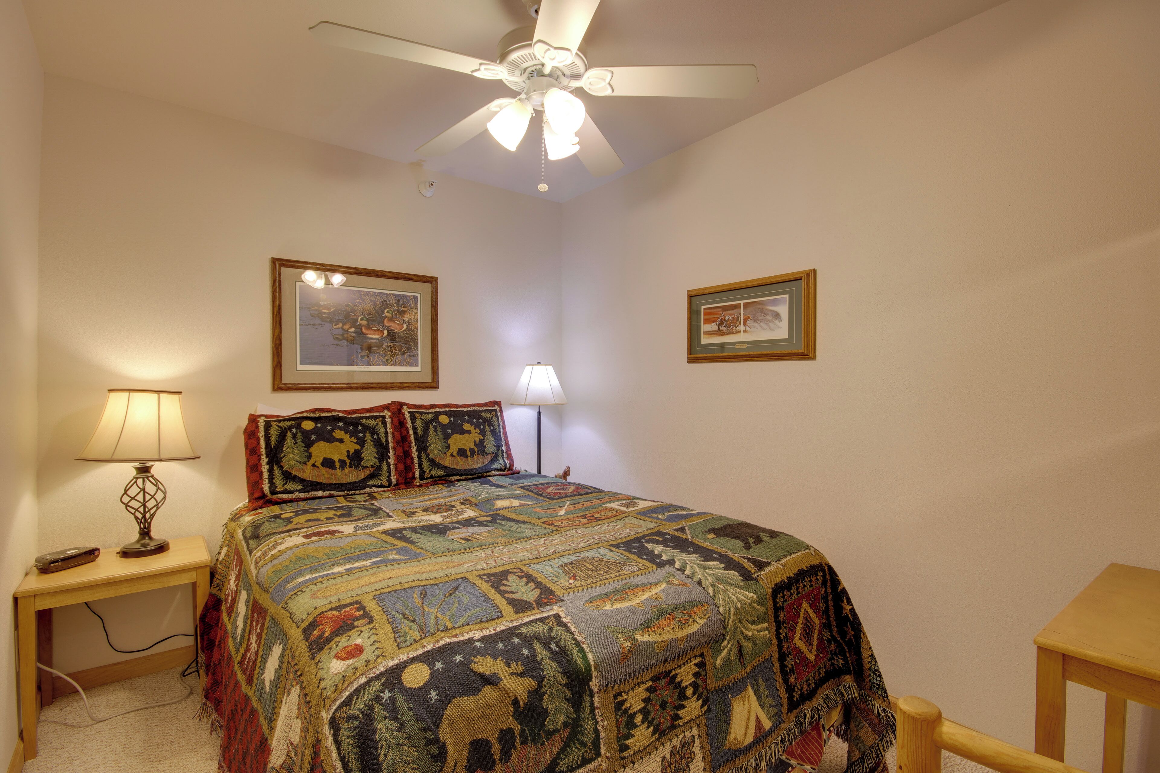 Une chambre à coucher avec un lit, un ventilateur de plafond, deux tableaux encadrés accrochés au mur, une lampe de chevet et une petite table.