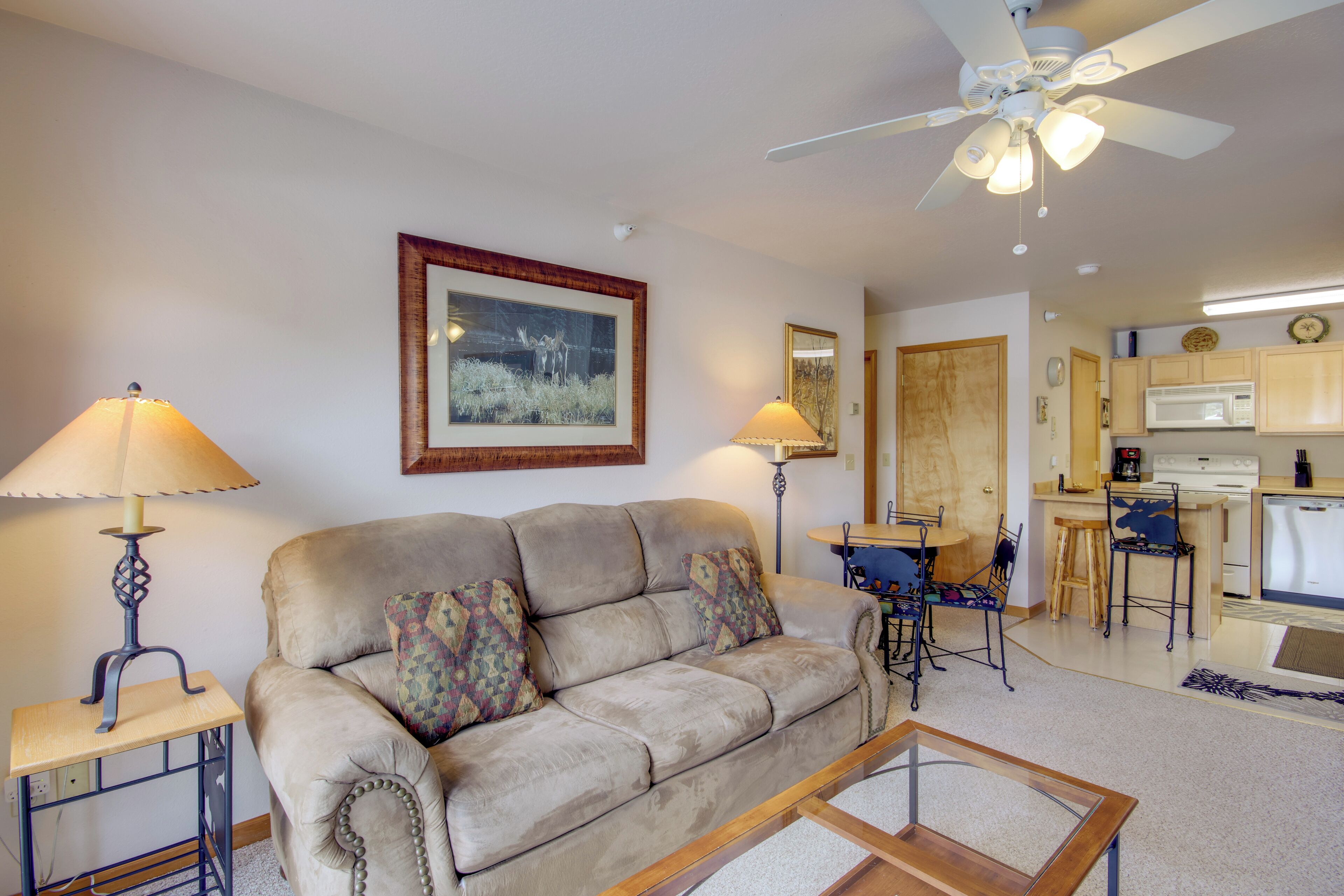 A living room with a sofa, two lamps, a ceiling fan, a dining area with chairs, and a kitchen with a microwave.