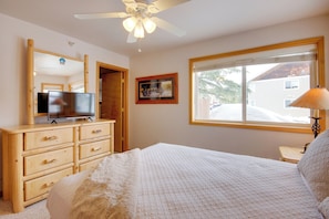 Une chambre à coucher avec un lit, une commode en bois, une télévision et une fenêtre donnant sur une maison.