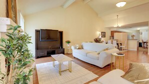 Uma sala de estar espaçosa com uma grande televisão de tela plana, um sofá branco, uma mesa de centro e uma área de refeições com cadeiras e uma pequena mesa.
