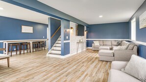 Amplia sala de estar con piso de madera, escalera, zona de bar y espacio de descanso con sofá y reposapiés.