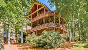 Een houten chalet met twee verdiepingen en een balkon, omgeven door bomen en een verzorgd gazon.