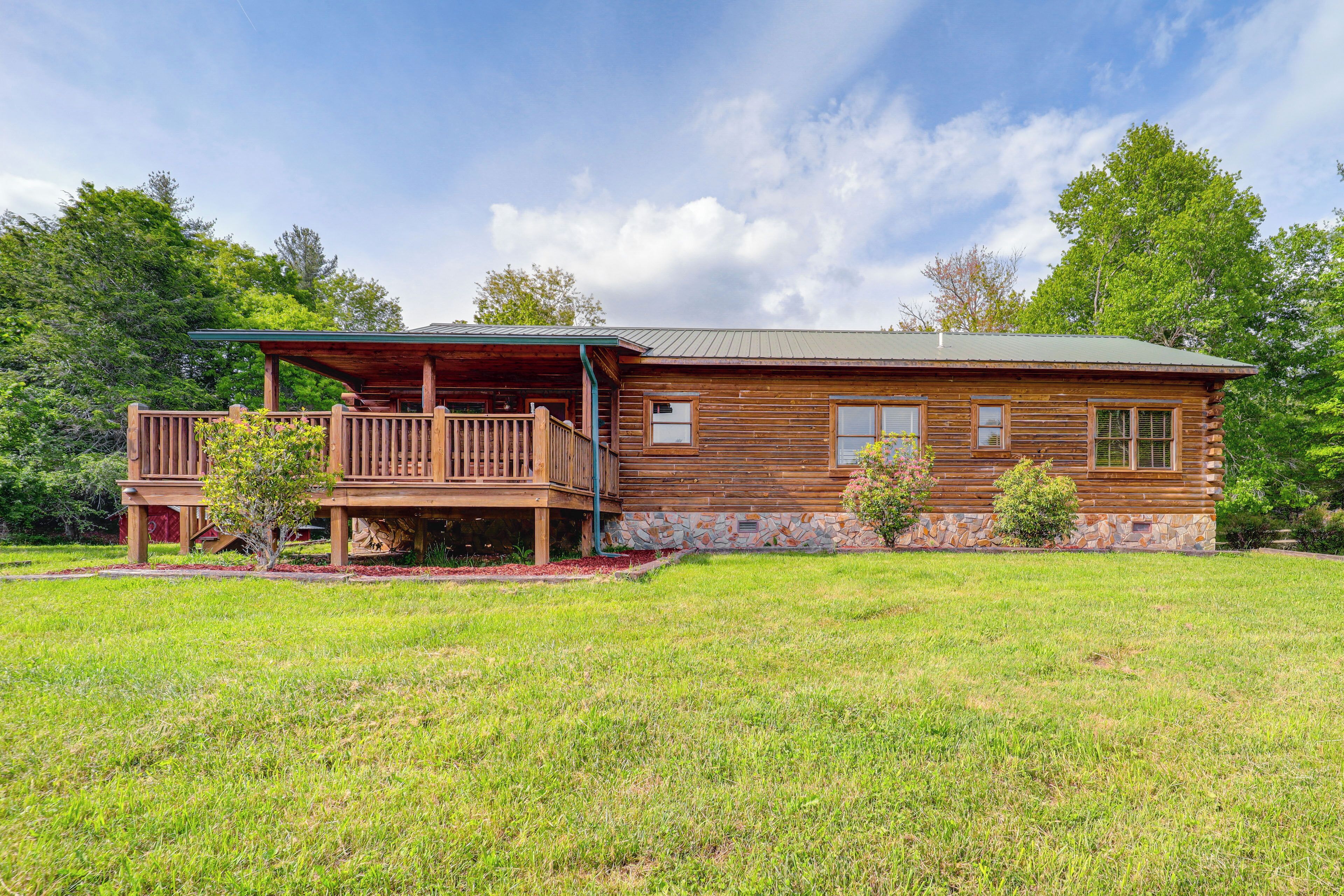 A wooden cabin with a porch and a stone foundation, surrounded by a grassy yard and trees.