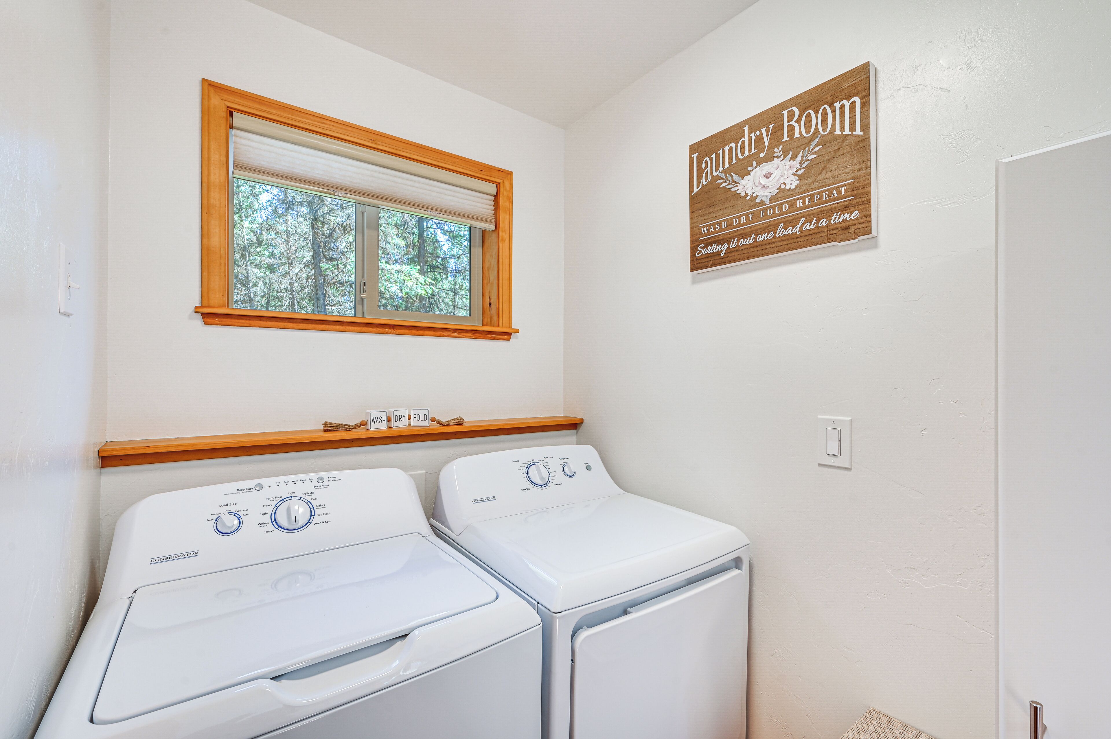 A laundry room with two front-loading washing machines, a window with a wooden frame, and a sign above the machines.