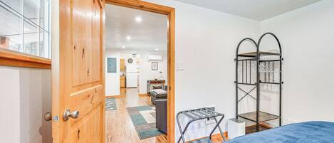 A room with a wooden door, a bed with a blue blanket, a metal drying rack, and a view into a kitchen area.