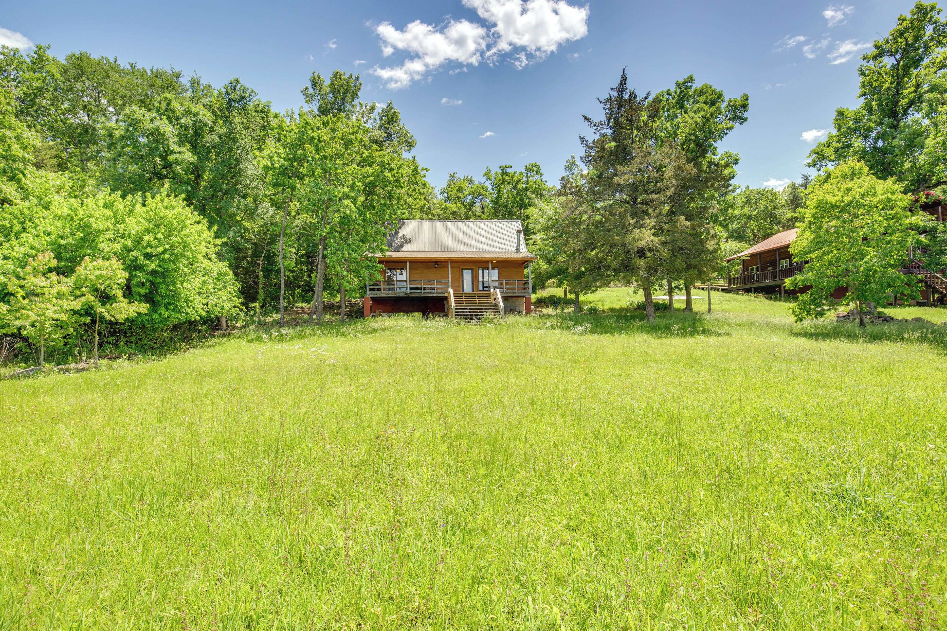 A cabin with a porch and a deck, surrounded by a grassy field and trees.