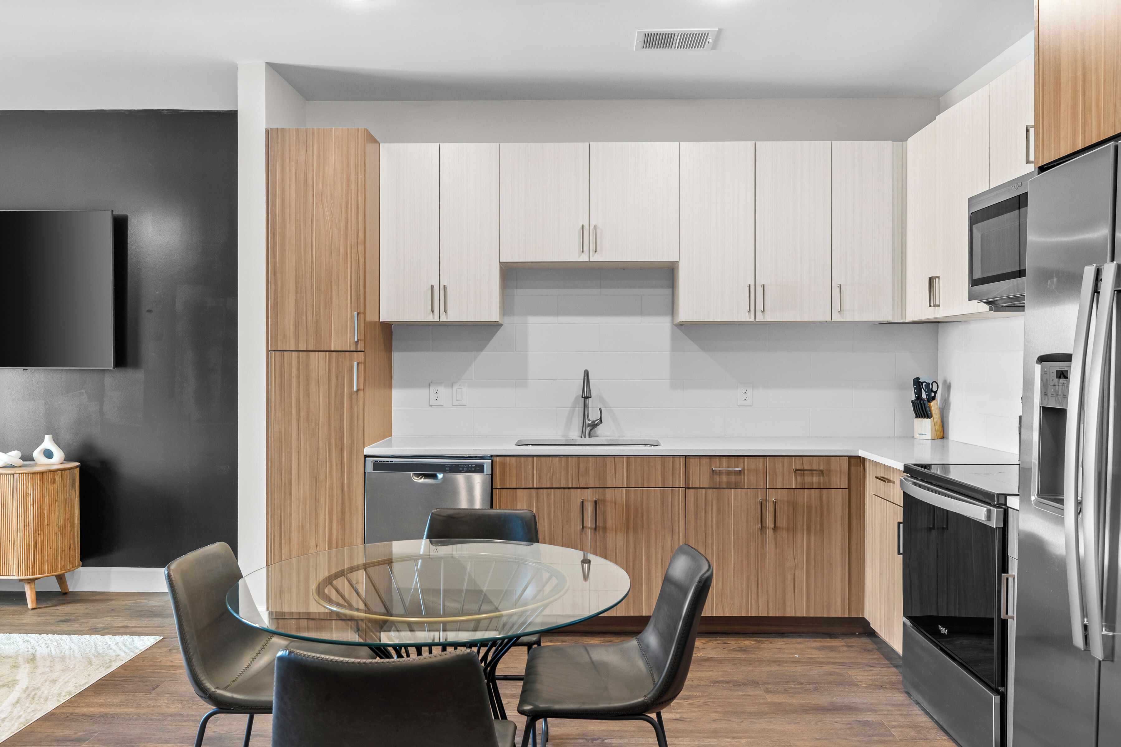 A modern kitchen with white cabinets, a stainless steel refrigerator, and a glass dining table with chairs.