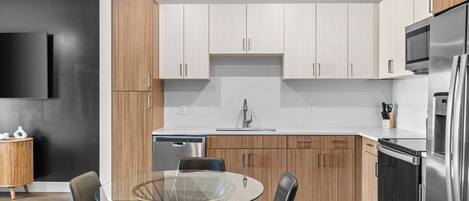 A modern kitchen with white cabinets, a stainless steel refrigerator, and a glass dining table with chairs.