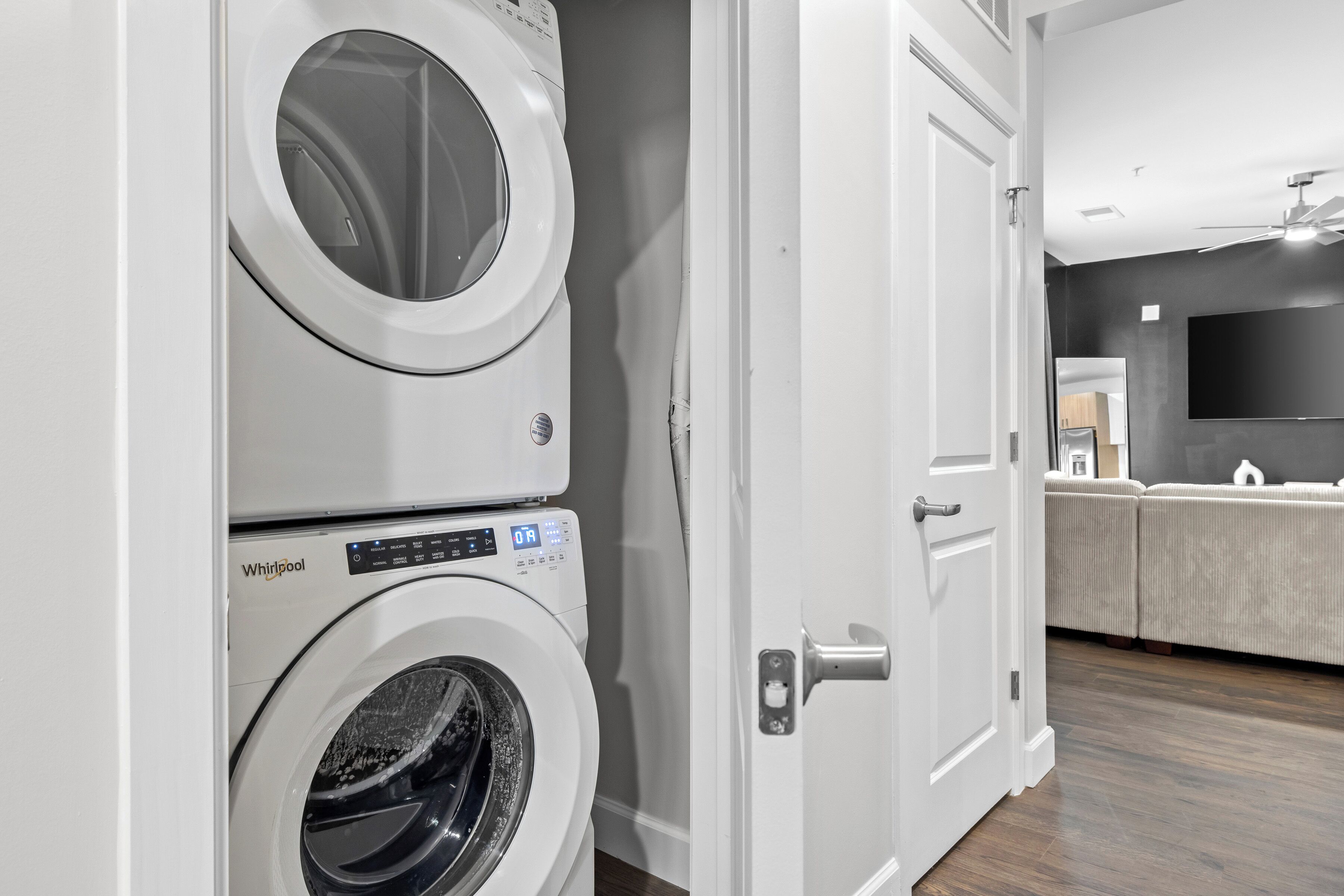 Uma lavanderia com duas máquinas de lavar Whirlpool, uma porta branca e um sofá cinza na área de estar adjacente.