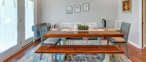 Comedor con una mesa de madera, sillas blancas y un banco. Hay tres láminas botánicas enmarcadas en la pared y una planta en maceta sobre la mesa.