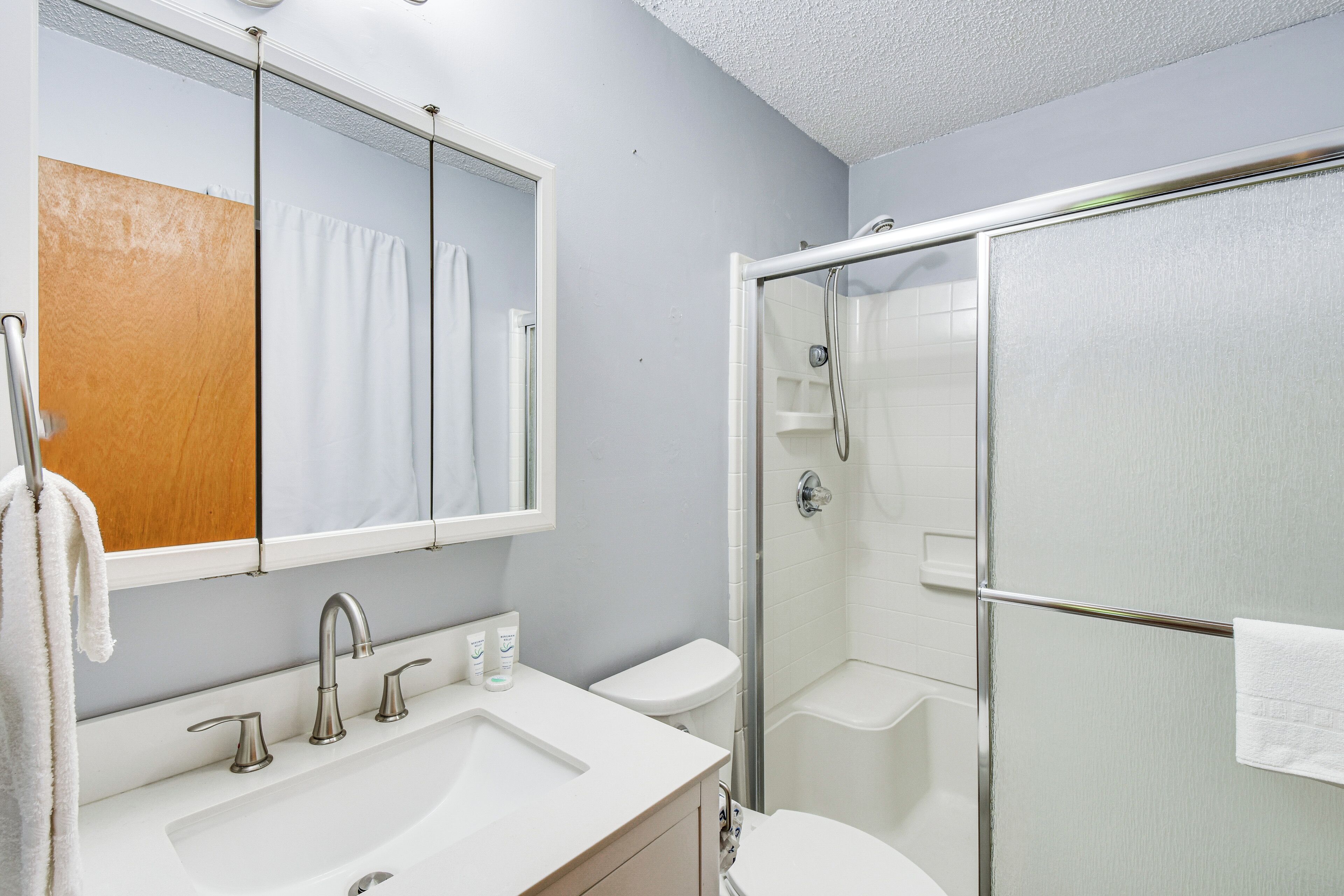 A bathroom with a white sink, mirror, and toilet, a glass shower enclosure, and a towel hanging on the wall.