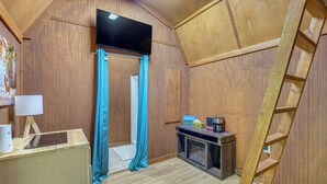 A wooden interior with a television mounted on the wall, a wooden ladder, a fireplace, and a table with a lamp and a speaker.