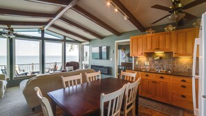 A dining area with a wooden table and white chairs, a kitchen with wooden cabinets, and a view of the sea through large windows. - Dreamy Lake Poinsett Cabin w/ Deck, Dock & Views! (Arlington)