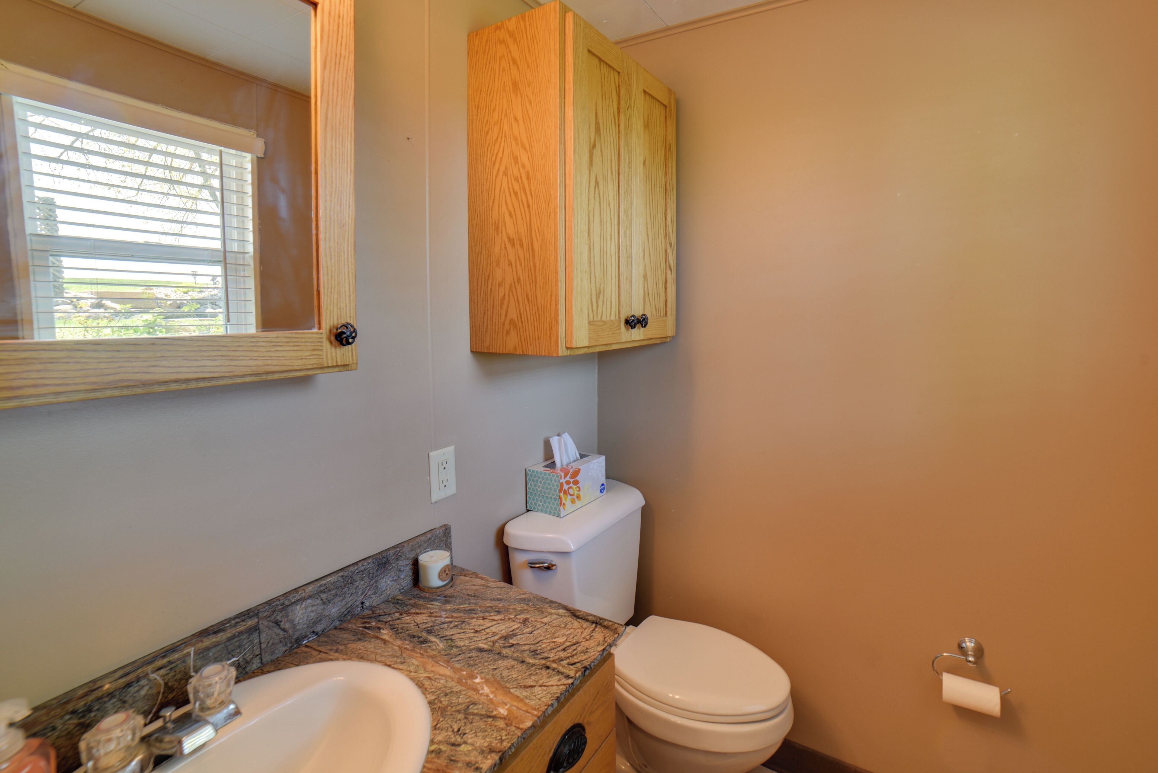 A bathroom with a sink, toilet, wooden cabinet, and a window with blinds.