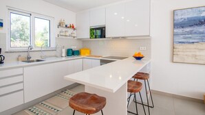 Een moderne keuken met witte kasten, een centraal eiland met krukken, een groot raam en een schilderij met een strandthema aan de muur.