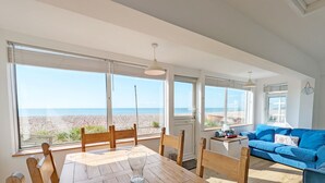 A dining area with a wooden table and chairs, large windows with a view of the beach, and a blue sofa in the living area.