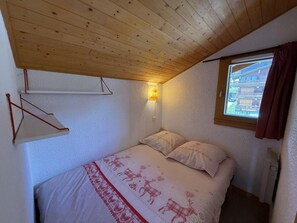 A bedroom with a wooden ceiling, a bed with a patterned bedspread, a wall-mounted light, and a window with curtains.
