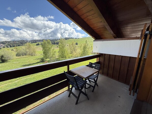 A balcony with a table and chairs, offering a view of a green landscape and distant mountains.