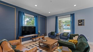 Una sala de estar con un televisor de pantalla plana grande, una mesa de centro de madera, un sofá y un sillón. Hay dos ventanas con cortinas azules y un cuadro en la pared.