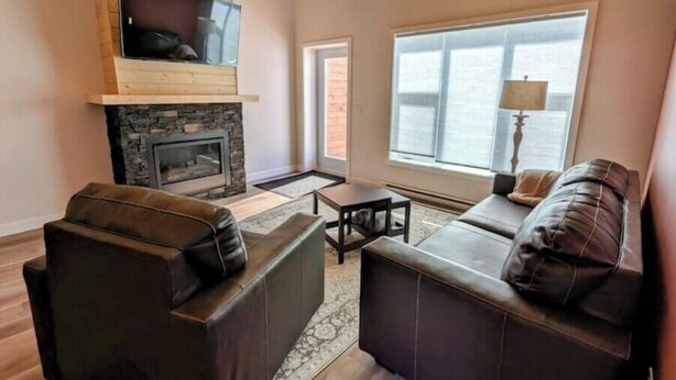 A living room with a stone fireplace, wooden ceiling, and large windows. - Yukon Hot Springs Villa #33 (Whitehorse)