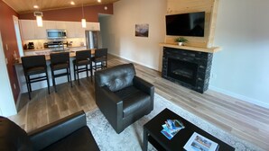 A modern living room with a fireplace, a dining area with a kitchen in the background, and a flat-screen TV mounted above the fireplace.