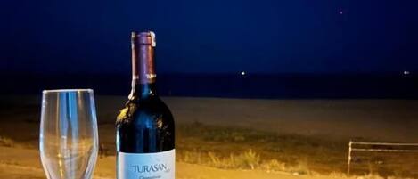 A bottle of Turasan wine and an empty glass on a table outside at night.