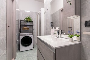 A modern bathroom with grey tiles, a built-in washing machine, a sink with a mirror, and a shelf with a plant and toiletries. - Il Rifugio del Viaggiatore by Wonderful Italy (Torino)
