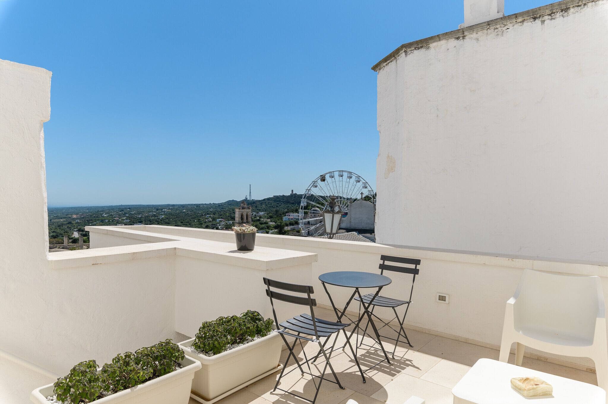Une terrasse sur le toit, avec un mur blanc, deux tables accompagnées de chaises, et une vue sur une grande roue et un paysage lointain.