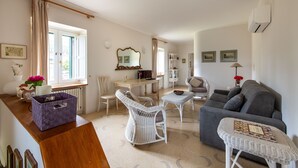 A spacious living area with a grey sofa, wicker chairs, a small table, and a dining table with chairs. There is a window with curtains, a radiator, and a wall-mounted air conditioner. - Dependance a Baglio il Corvo by Wonderful Italy (Gangi)