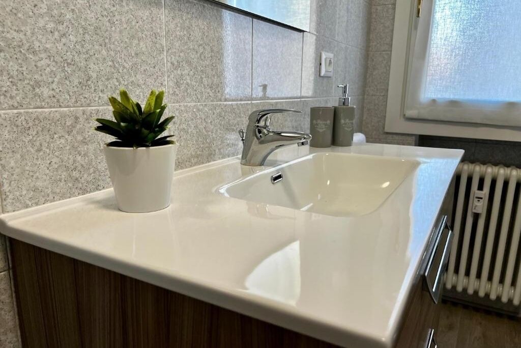 Un baño con lavamanos blanco, grifo moderno, planta en maceta y ventana con persianas.