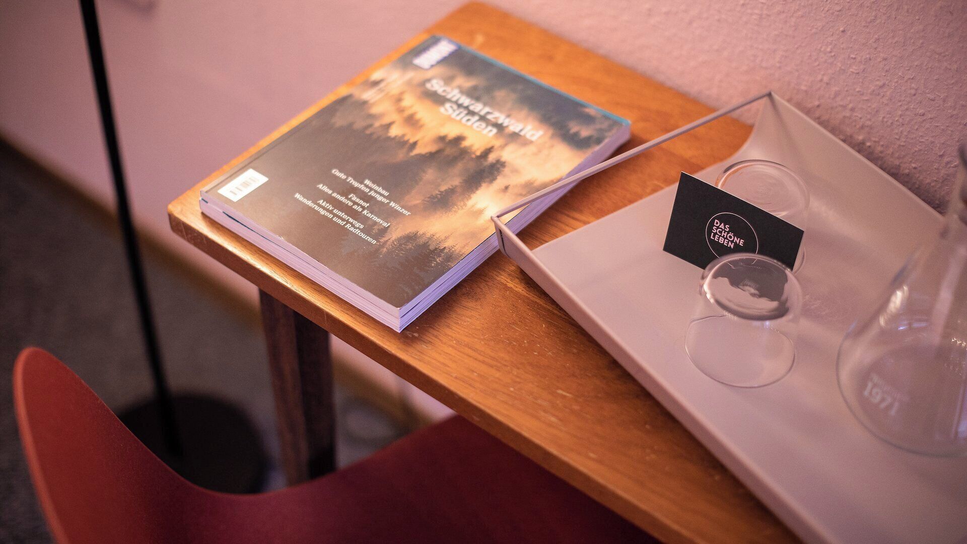 A book titled 'Schwarzwald Sieden' on a wooden table, a glass with a label, and a red chair.