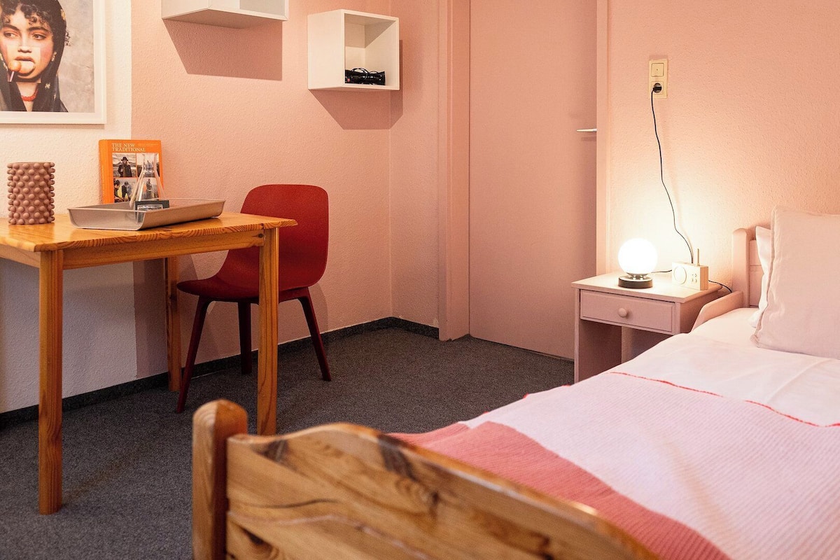 Une chambre d’hôtel avec un bureau en bois, une chaise rouge, une table de chevet avec une lampe et un lit avec un oreiller blanc.